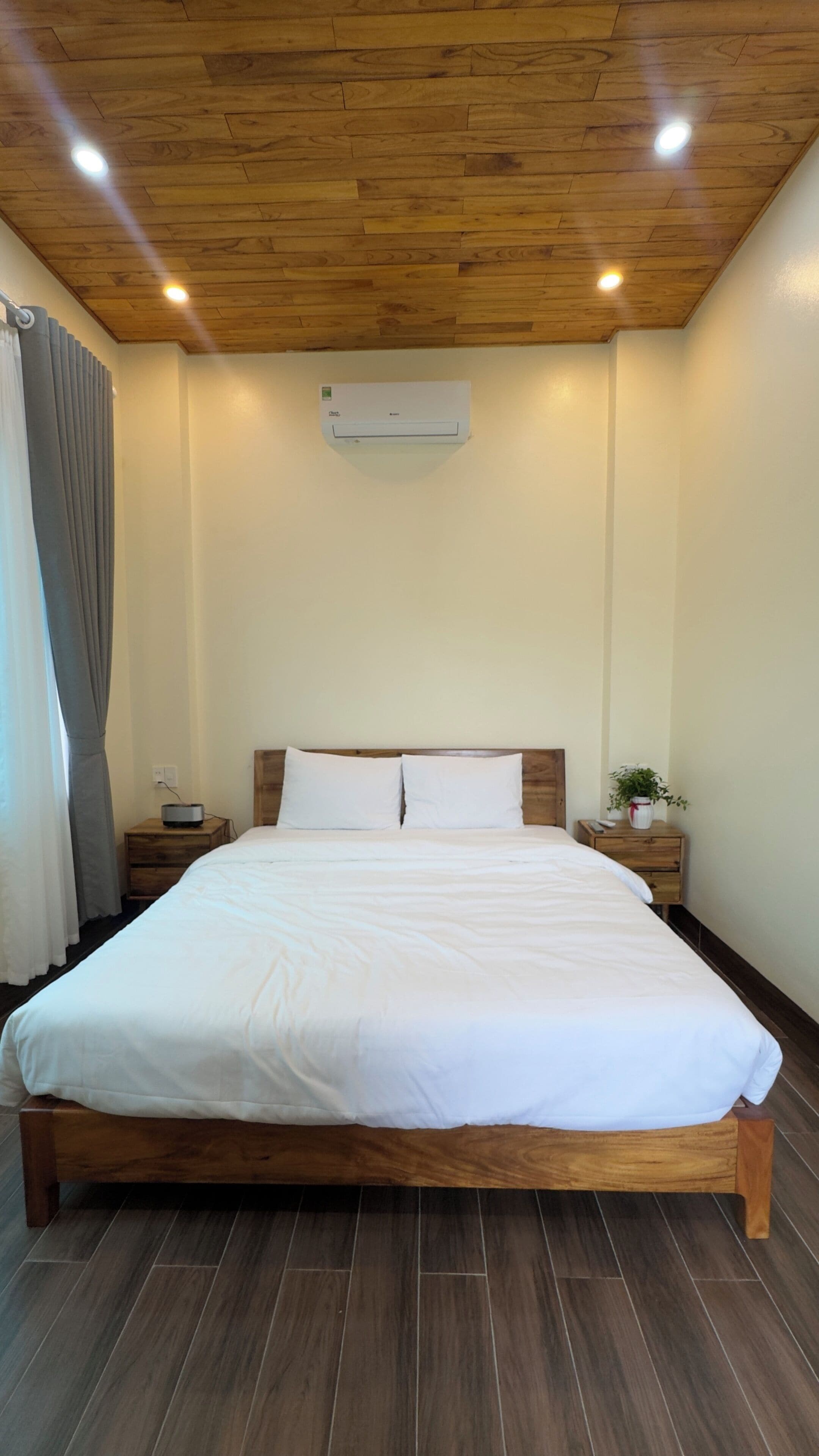 A bedroom with a wooden ceiling, a bed with white bedding, a nightstand with a lamp, and an air conditioning unit on the wall.