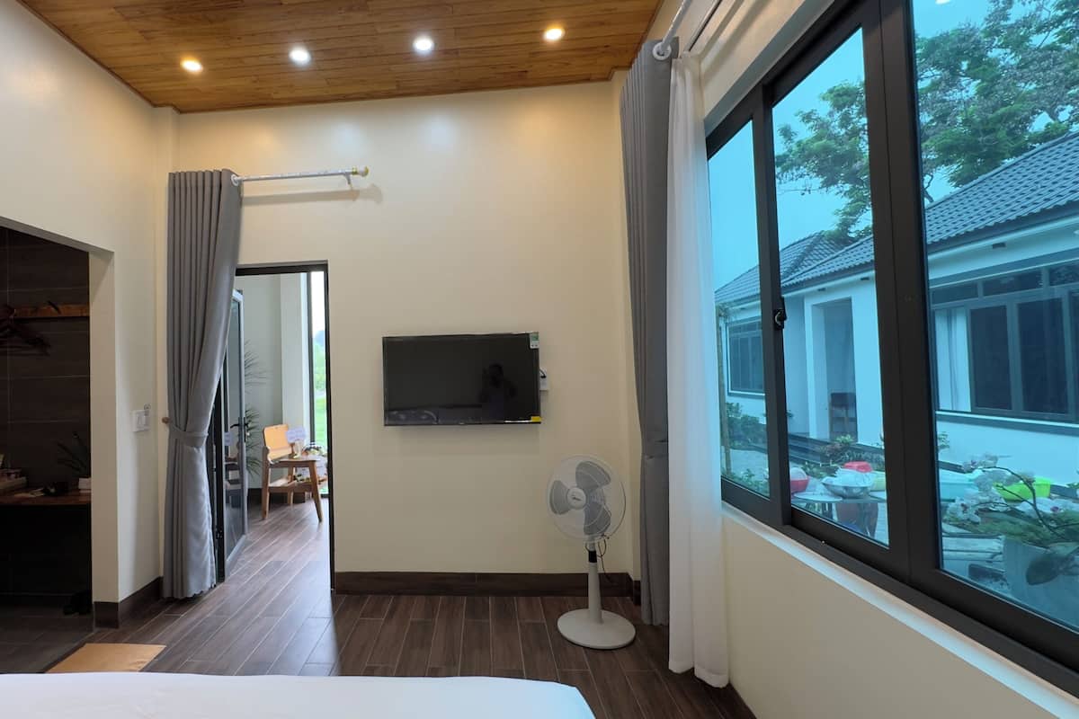 Un dormitorio con un ventanal grande, un televisor de pantalla plana, un ventilador de pie y techo de madera.