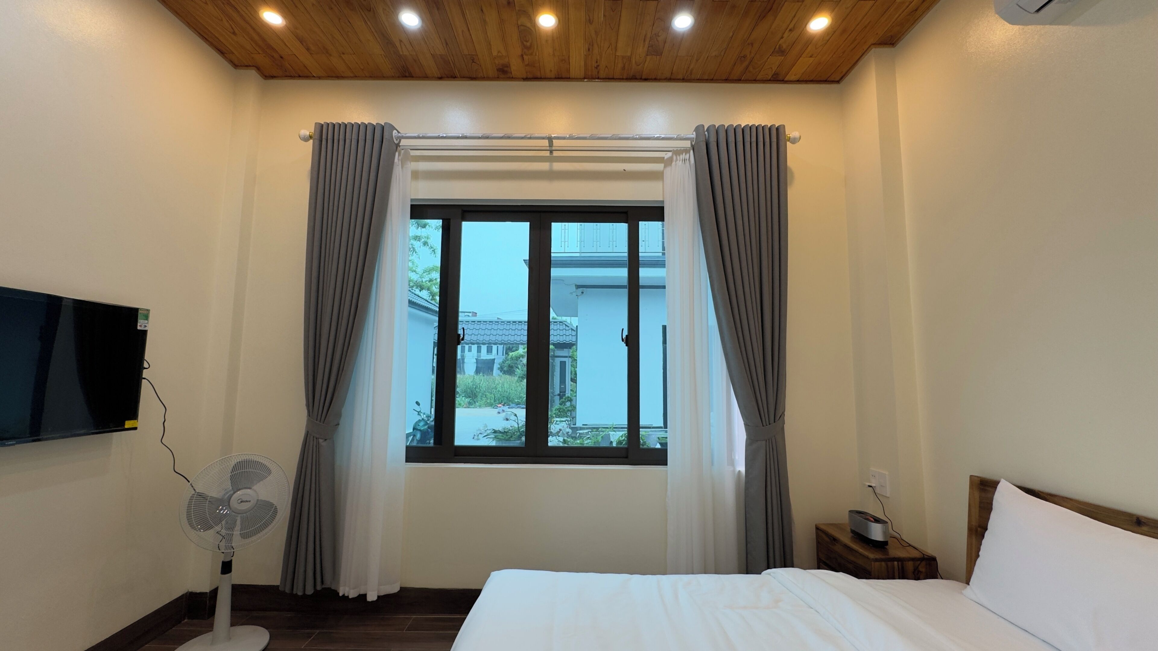 A hotel room with a bed, a TV mounted on the wall, a window with curtains, a fan, and a bedside table with a lamp.