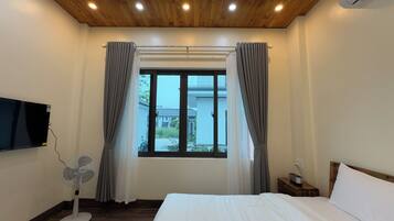 A hotel room with a bed, a TV mounted on the wall, a window with curtains, a fan, and a bedside table with a lamp.