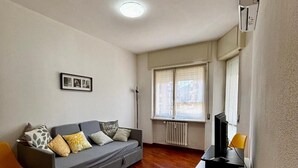 Un salon avec un canapé gris, une petite table sur laquelle se trouve un téléviseur, un radiateur et une fenêtre avec des stores.