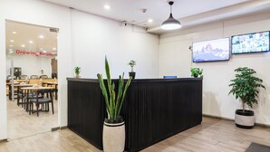 A reception area with a black counter, potted plants, a TV on the wall, and a drawing room visible through a glass door.