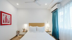 A hotel room with a bed, a ceiling fan, an air conditioning unit, a nightstand with a lamp, a small plant, and a framed picture on the wall.