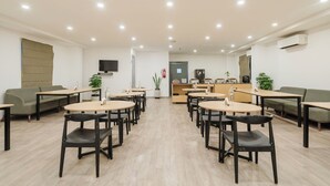 A modern, well-lit room with round tables and chairs, a sofa, and a reception desk with a television.