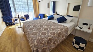 Quarto de hotel moderno com uma cama grande, área de estar com mesa e cadeiras, televisão e minibar.