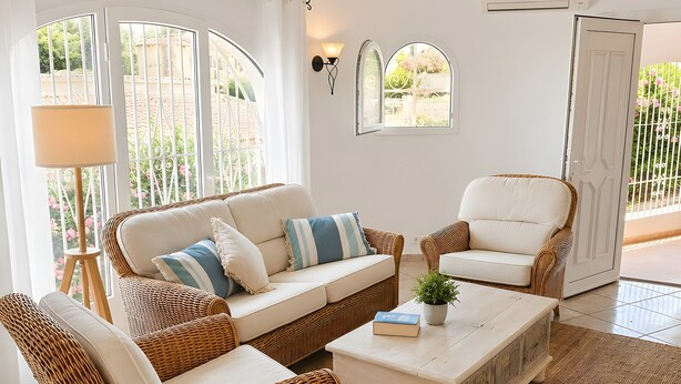 Ein Zimmer mit weißer Decke, Korbmöbeln, einem weißen Sofa, einem weißen Sessel und einem weißen Couchtisch, mit Blick auf Grünfläche.