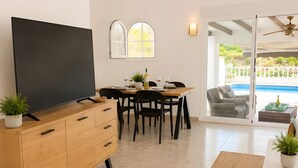 Ein modernes Wohnzimmer mit Flachbildfernseher, Holzschrank, Esstisch, Stühlen und Blick auf einen Pool durch Glastüren.