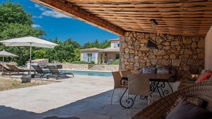 Ein überdachter Außenbereich mit einer Steinwand, einem Pool, Liegestühlen und einem Tisch mit Stühlen.