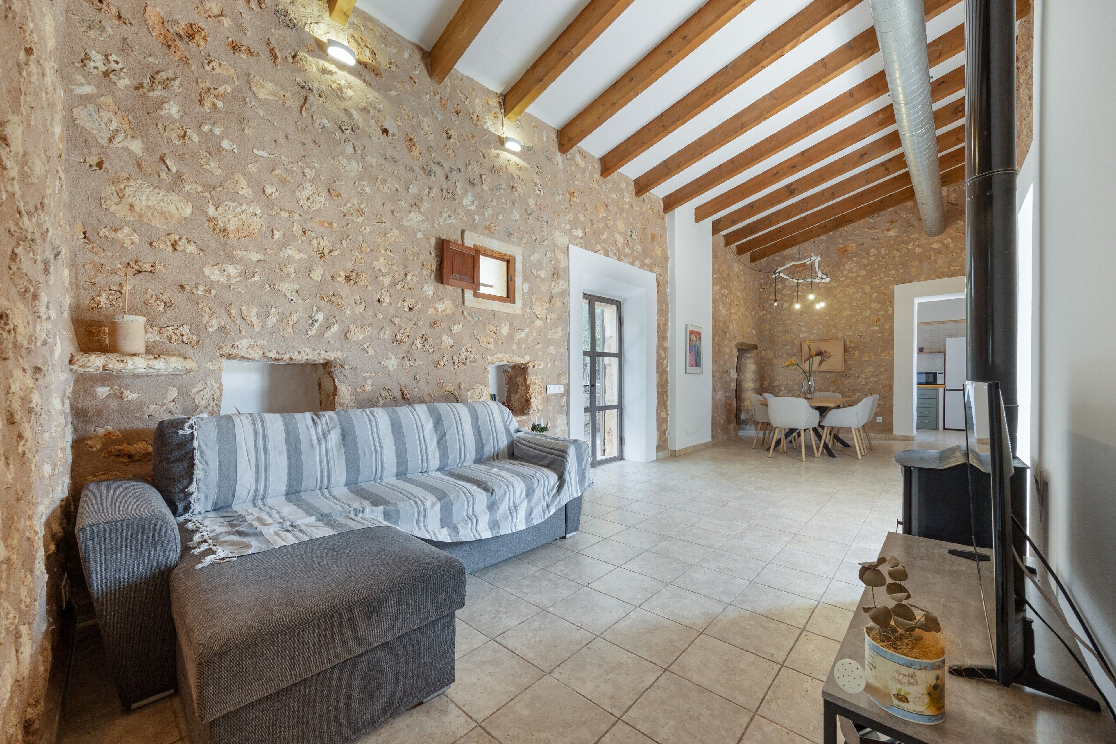 Una sala de estar con pared de piedra, vigas de madera, un sofá, una mesa de centro y un área de comedor al fondo.