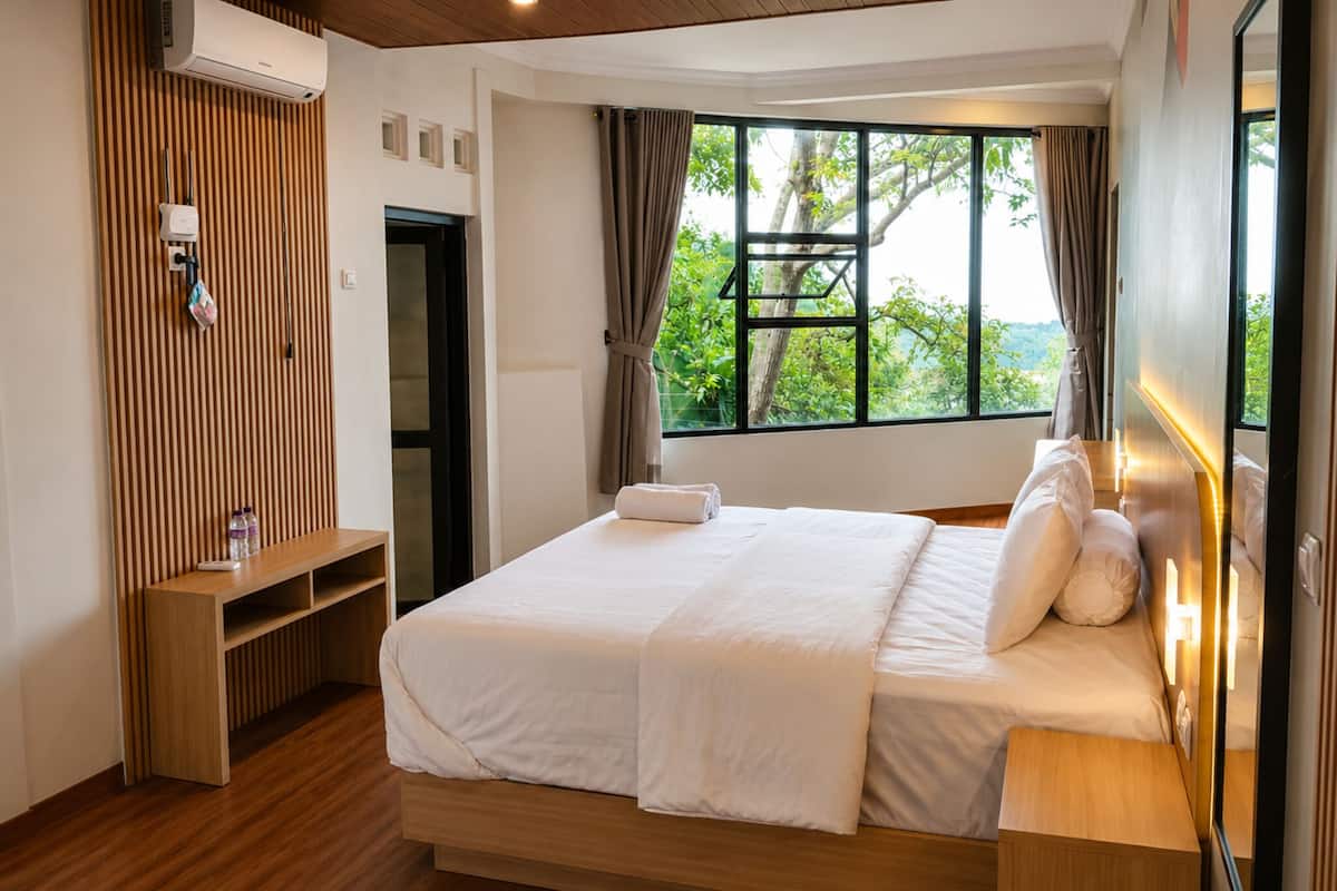 Habitación de hotel con cama, repisa de madera, ventana con cortinas y vista a vegetación.