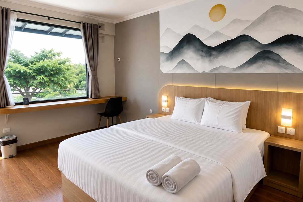 A hotel room with a large bed, a view of greenery outside, and a painting of mountains on the wall.