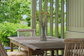Et træbord med en klar glasvase med tørrede blomster, placeret på en veranda med træstole.