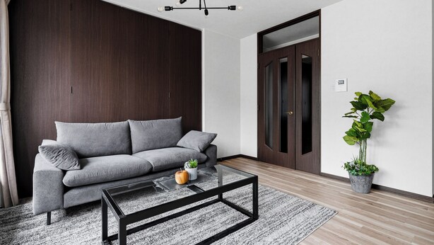 Una sala de estar moderna con un sofá gris, una mesa de centro de vidrio, piso de madera, una planta grande en maceta y una puerta de madera oscura.