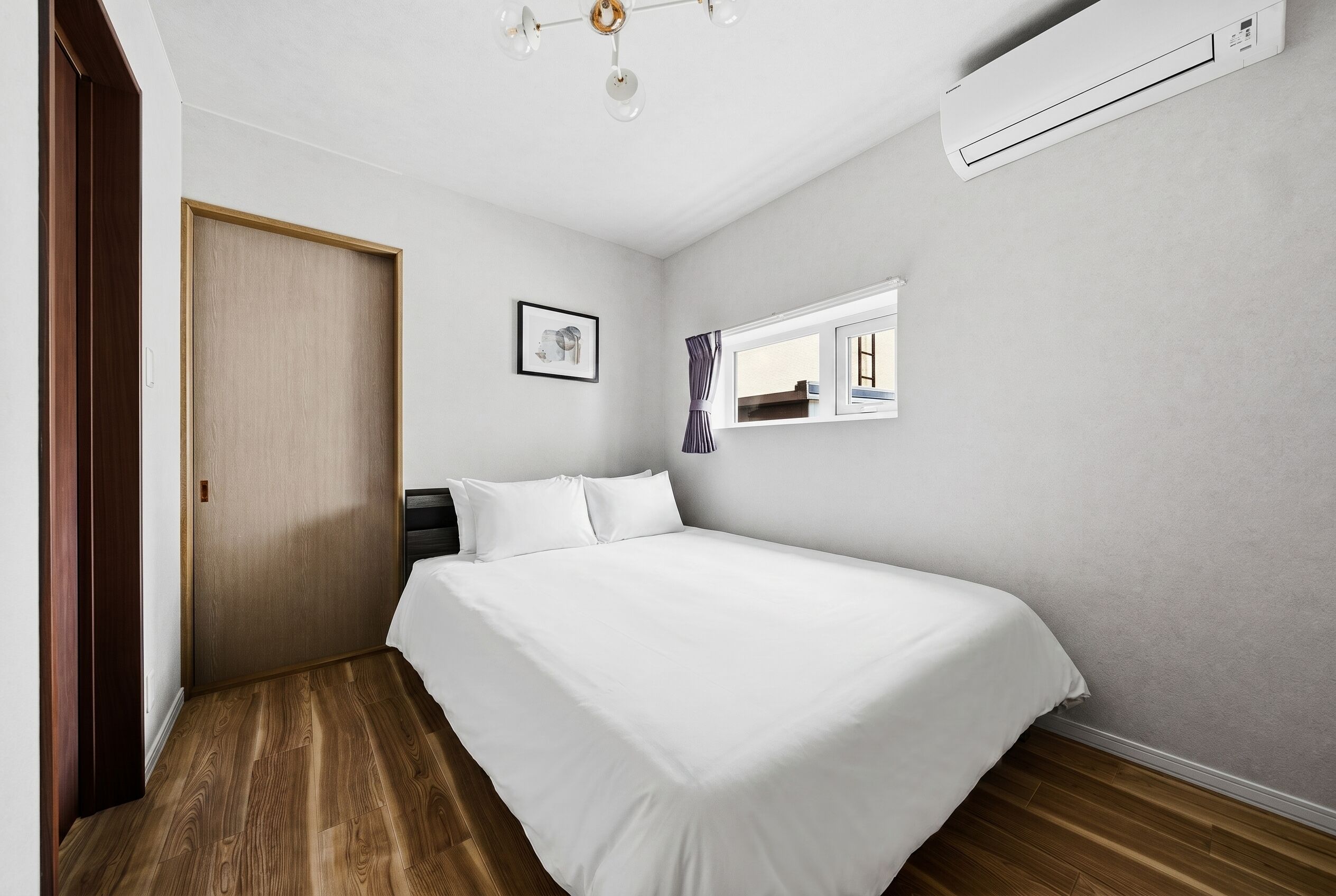 A bedroom with a bed, wooden flooring, a window with curtains, a door, and an air conditioning unit.