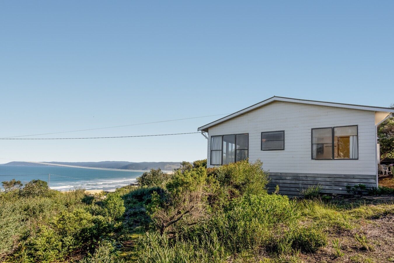 Casa de una planta con fachada blanca, ubicada en una colina cubierta de césped con vistas a un paisaje costero bajo un cielo despejado.