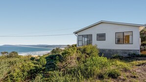 Une maison de plain-pied à l’extérieur blanc, située sur une colline verdoyante qui offre une vue sur un paysage côtier sous un ciel dégagé.