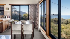 Un espace repas avec une table et des chaises, une cuisine avec un comptoir et des appareils électroménagers, et de grandes fenêtres offrant une vue sur l’extérieur.