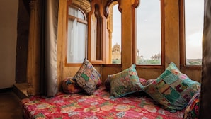 Una habitación con una cama, almohadas coloridas y vistas a un edificio lejano a través de ventanas arqueadas.