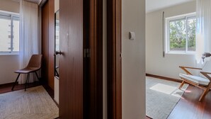 Ein Zimmer mit Holztür, einem Stuhl, einem Fenster und Blick ins Grüne.