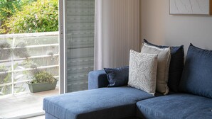 Ein modernes Wohnzimmer mit einem grauen Ecksofa, einer Topfpflanze und einem großen Fenster mit Jalousien.