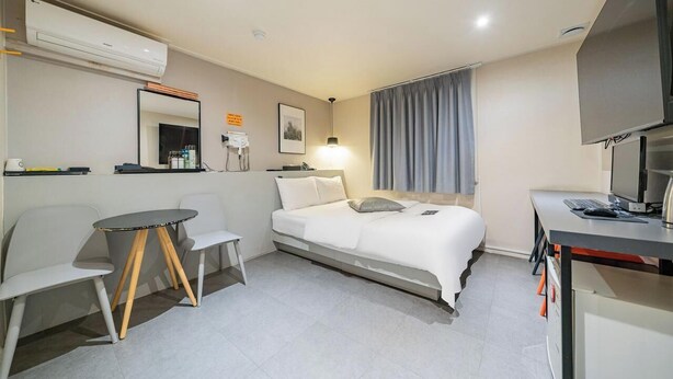 A hotel room with a bed, a desk with a computer, a chair, a small table, a mirror, an air conditioner, and a window with curtains.