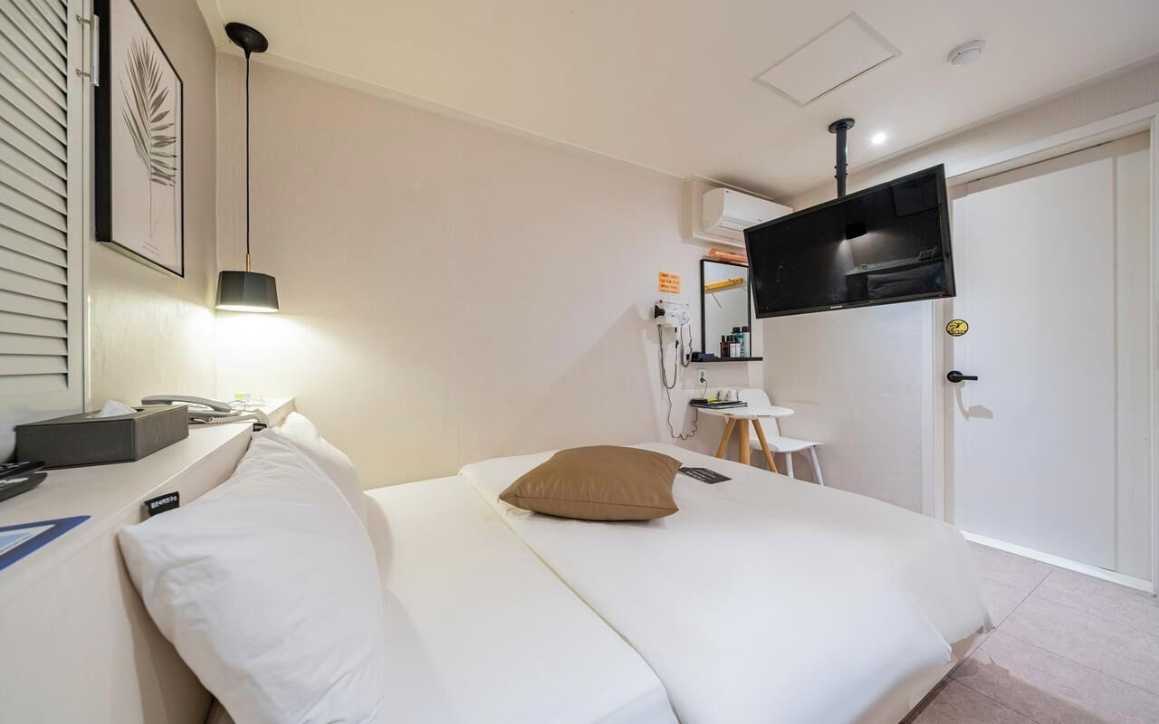 A hotel room with a bed, a desk with a telephone, a television mounted on the wall, and a door with a lock.