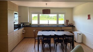A modern kitchen with wooden cabinets, a dining table with chairs, a window with a view of greenery, and a white ceiling. - A very pleasant house with a beautiful garden, located 500 m from the harbor and the beach in Le Crotoy (Le Crotoy)