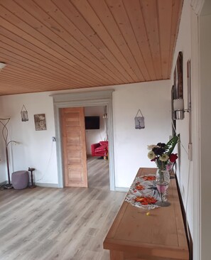 A room with a wooden ceiling, a wooden floor, a table with a floral tablecloth, a vase with flowers, and a red chair in the background.