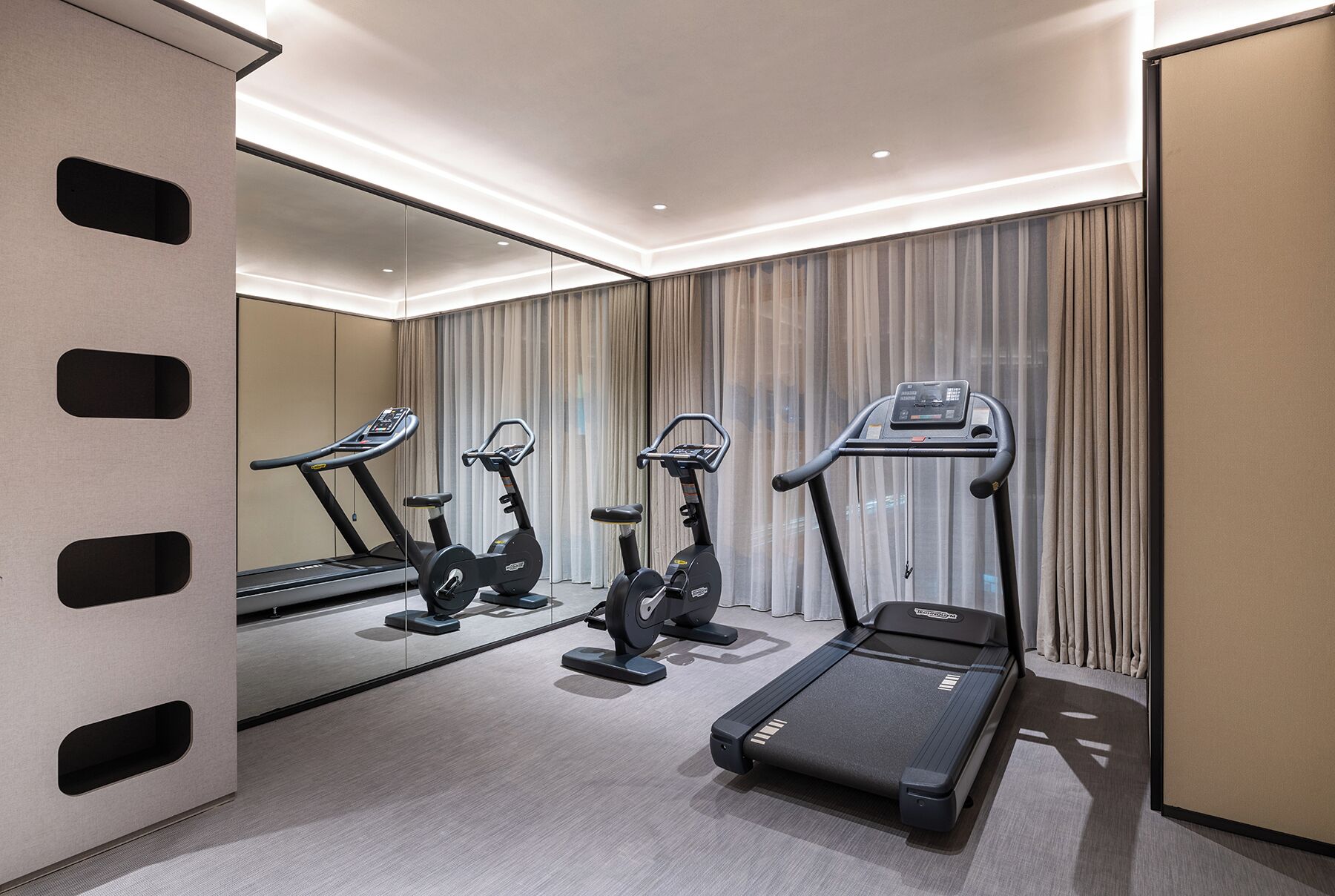 A hotel room with a treadmill, elliptical, and stationary bike, a large mirror, and a view of the city through the window.