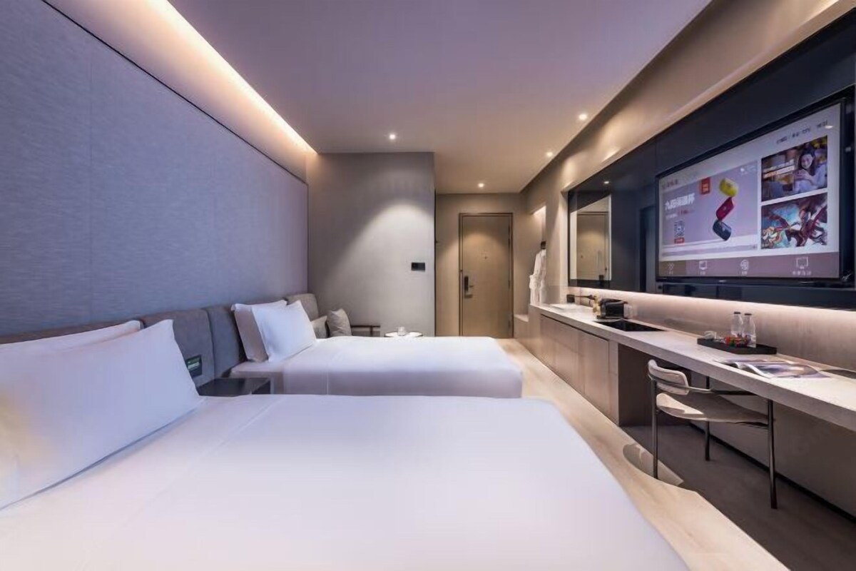 A modern hotel room with two beds, a desk, a large TV screen, and a bathroom door.