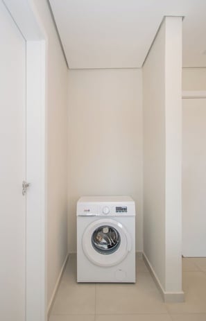 A compact, white, wall-mounted washing machine in a small, neutral-colored room with a door on the left and a white pillar on the right. - Corporate Stays Zuba 10 (Luque)