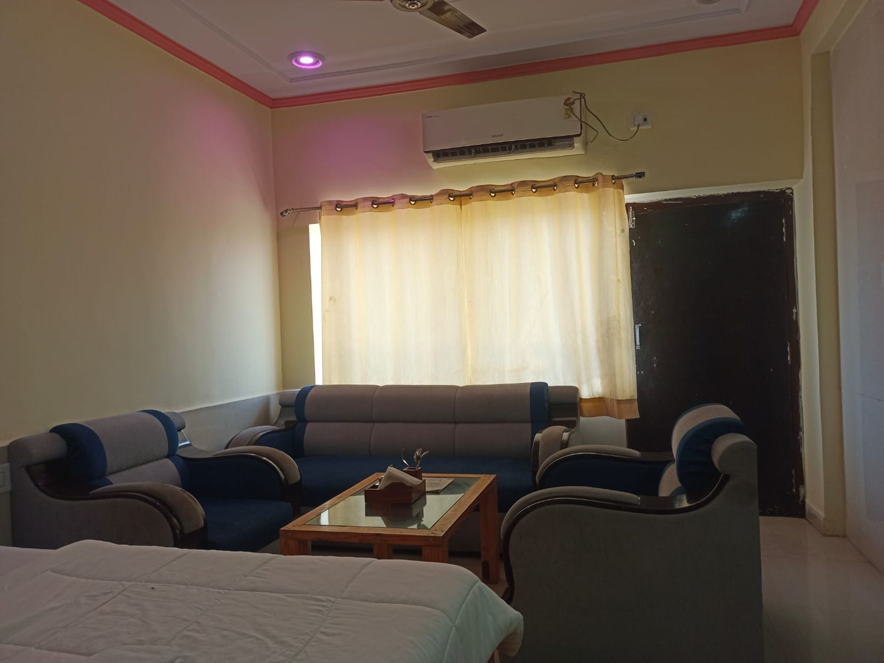 Una habitación con un sofá, dos sillones, una mesita, una cama, un aire acondicionado y una puerta.