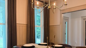 A dining room with a long wooden table set for four, a chandelier, and a view of the outdoors through large windows.