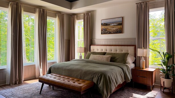A bedroom with a large bed, a bench, a nightstand, a lamp, and a potted plant.