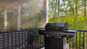 A barbecue grill on a wooden deck with a railing, surrounded by greenery.