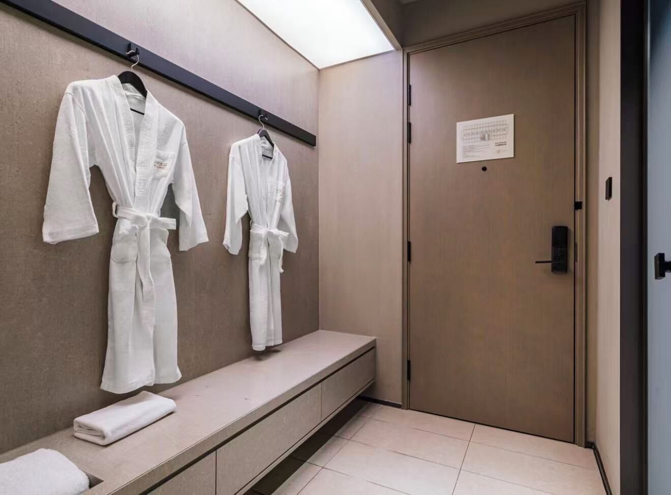 A hotel bathroom with two white robes hanging on a wall, a bench with a towel, and a door with a sign.