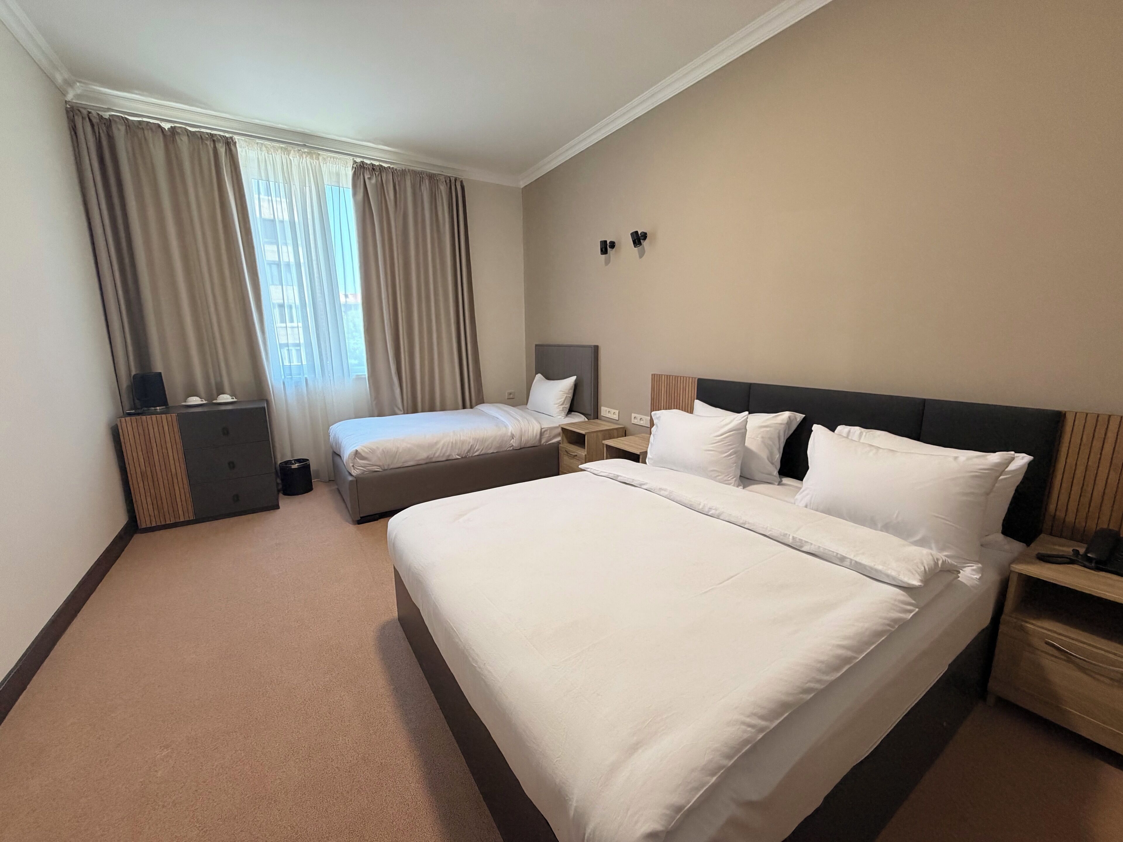 Habitación de hotel con una cama grande, una cama más pequeña, una mesita de noche, un tocador y un armario, además de una ventana con cortinas.