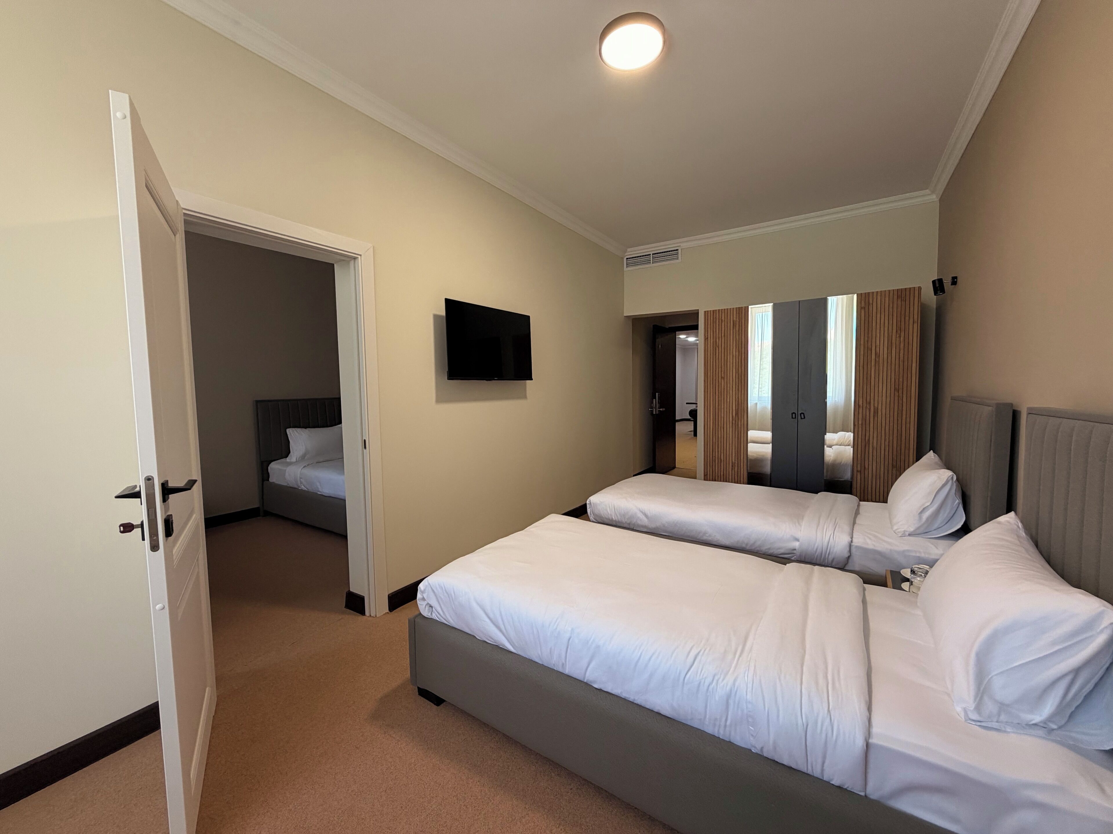 A hotel room with two beds, a television, a door leading to another room, and a ceiling light.