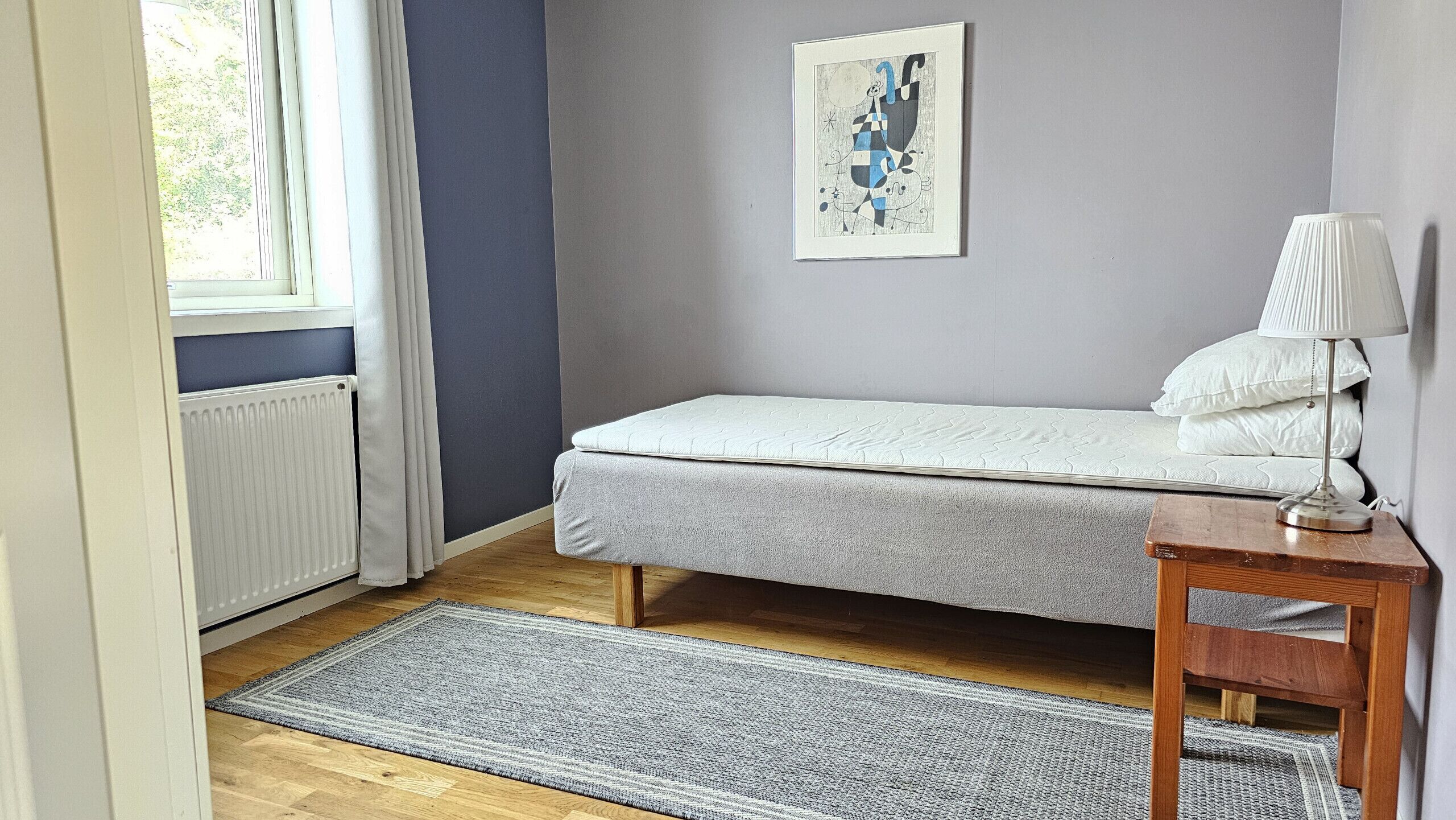 A bedroom with a bed, a bedside table with a lamp, a radiator, a window with curtains, and a painting on the wall.