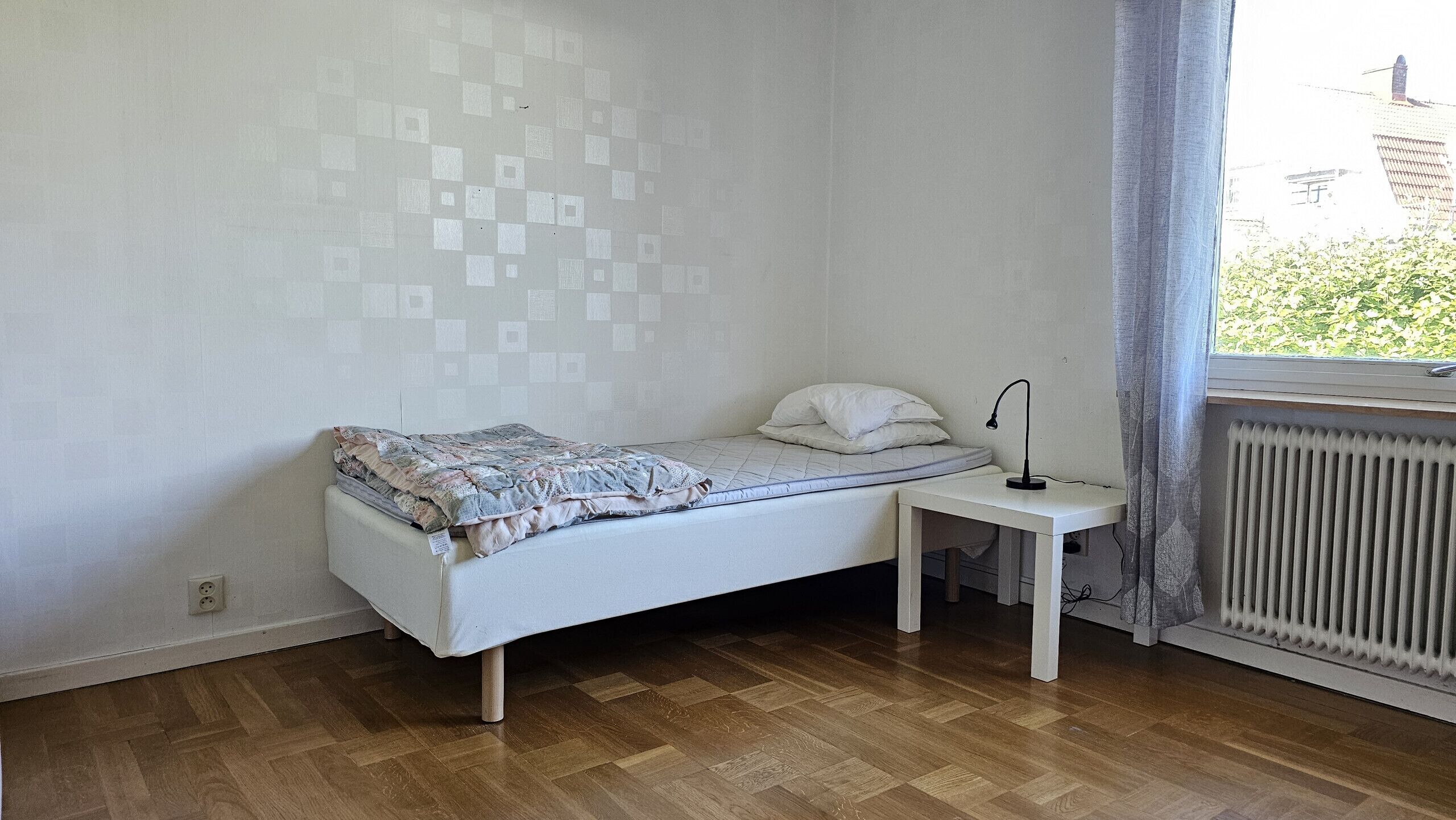 A small, single-bed room with a wooden floor, a bedside table with a lamp, and a window with a view of greenery.