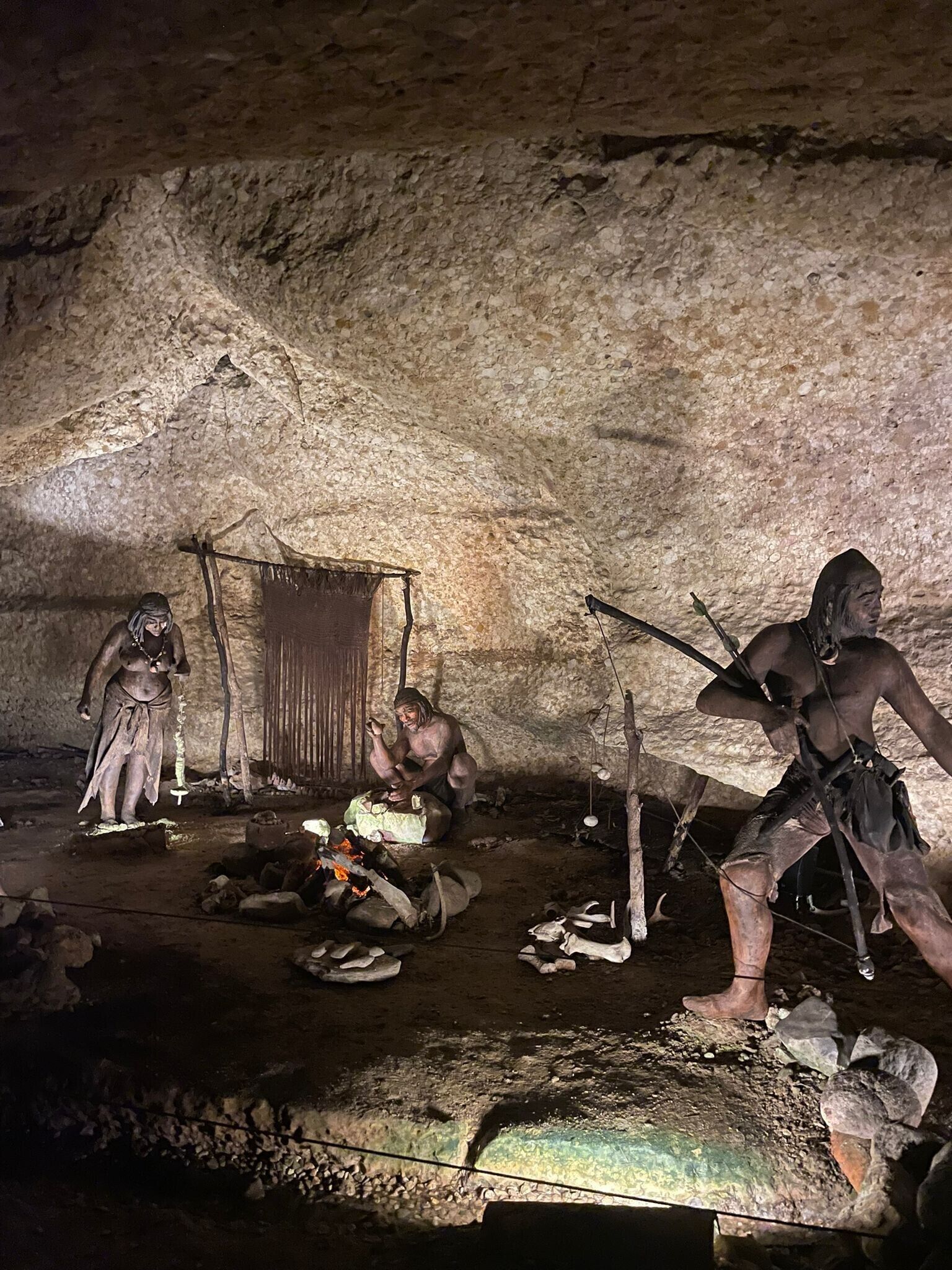 Interior de una cueva con figuras humanas prehistóricas, un hogar y herramientas de piedra.