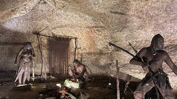 Interior de una cueva con figuras humanas prehistóricas, un hogar y herramientas de piedra.