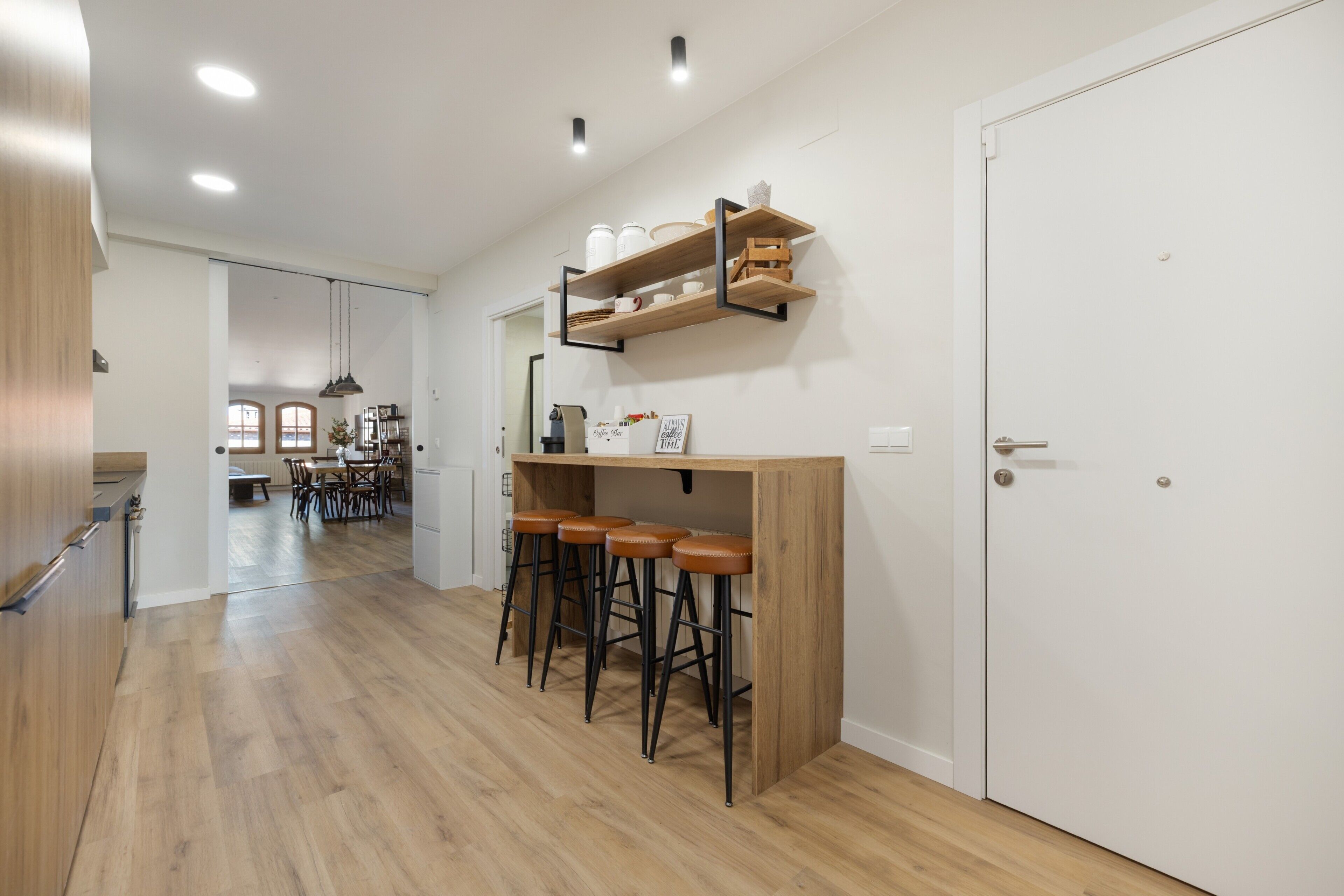 A modern kitchen with wooden bar stools, a built-in shelf with kitchenware, and a door leading to another room.