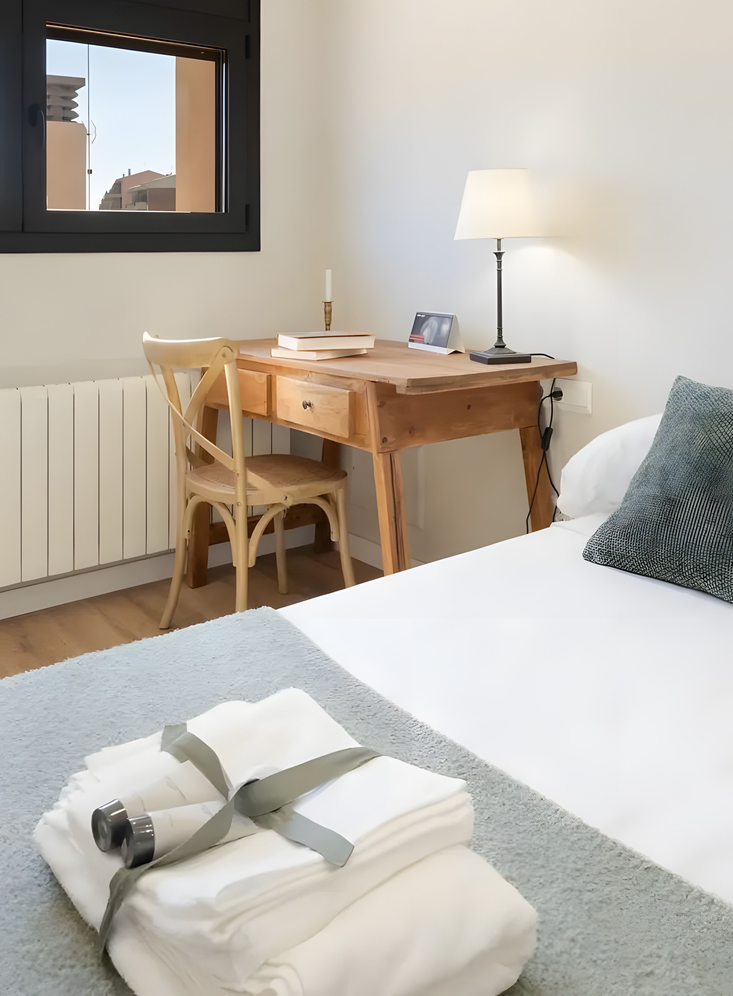 A hotel room with a bed, a wooden desk, a chair, a lamp, and a window with a view of buildings.