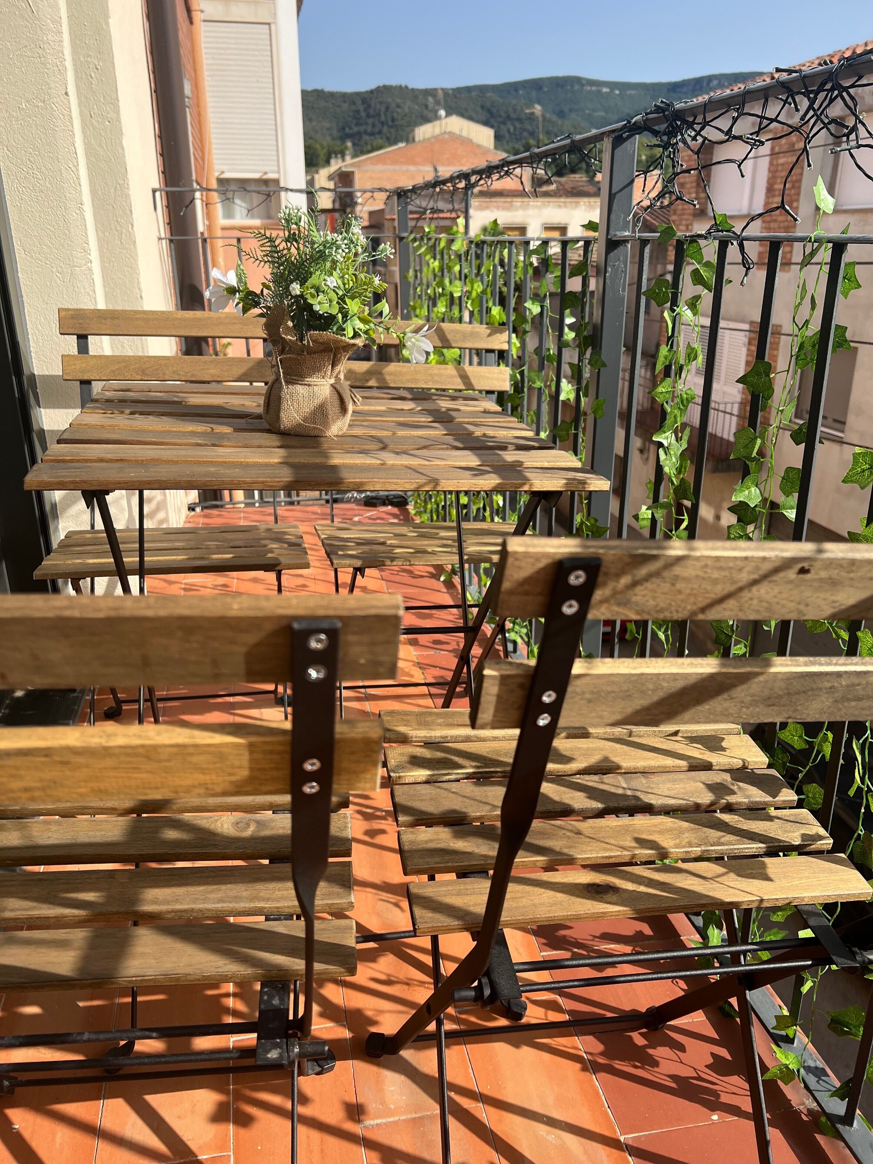 Una terraza al aire libre con mesas y sillas de madera, una planta en maceta y vistas a edificios y colinas al fondo.