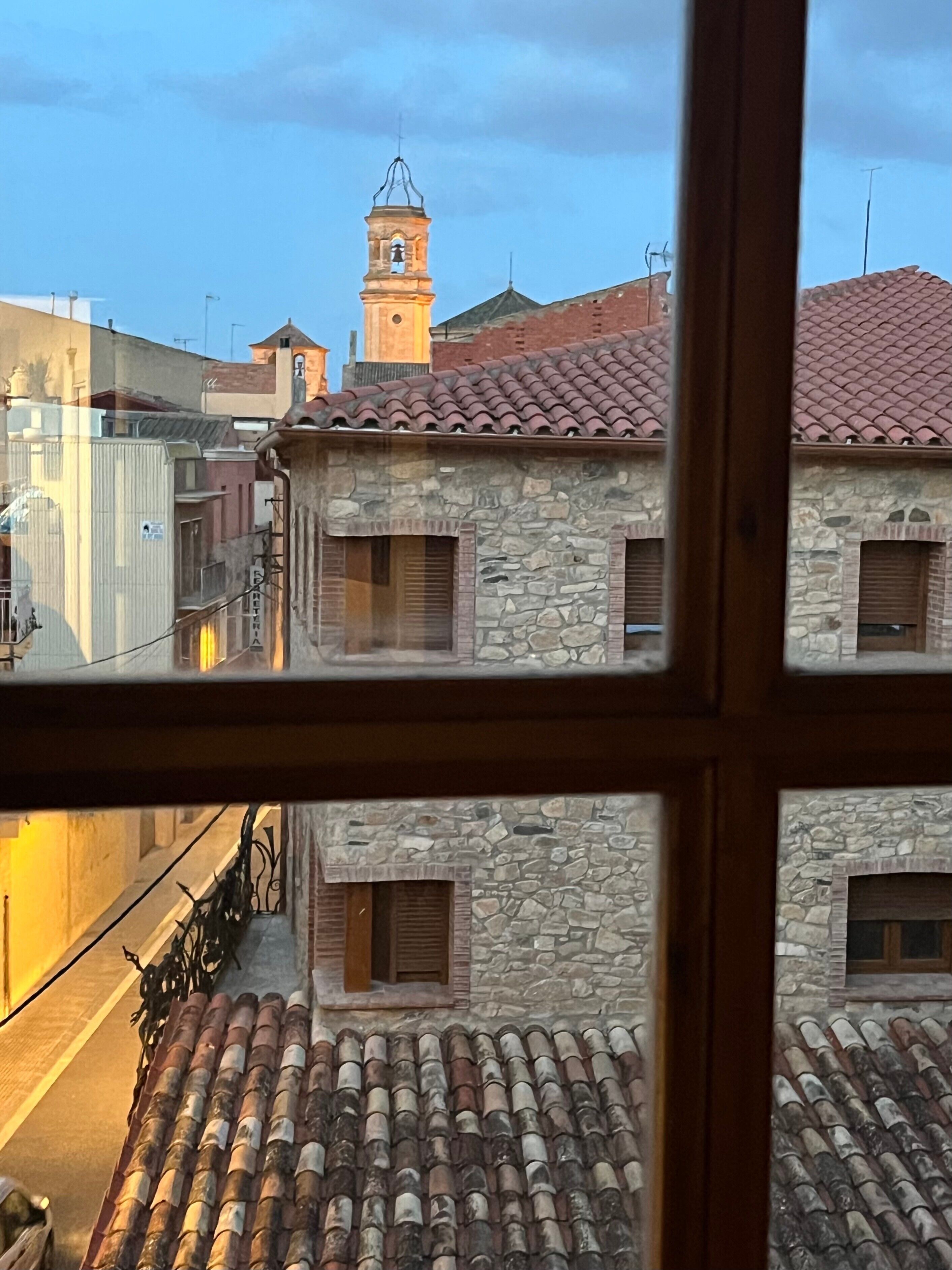 A view through a window of a building with stone walls, a tiled roof, and a bell tower.