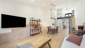 A spacious living area with a wooden bench, a white sofa, a TV mounted on a brick wall, and a staircase leading to an upper level.