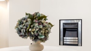 En vit vas med en blomsteruppsättning på ett runt bord, en modern öppen spis med en metallisk yta och en enkel, vit vägg.