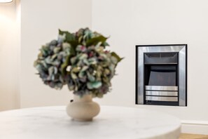 A white vase with a floral arrangement on a round table, a modern fireplace with a metallic finish, and a plain white wall. - Stylish Holland Park Patio Apartment (London)