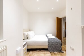 A small, modern bedroom with a bed, bedside table, and a door leading to another room. - Stylish Holland Park Patio Apartment (London)